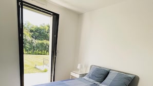 A bedroom with a large bed, a nightstand, a lamp, and a sliding glass door leading to a garden. - New house near Port-Manec'h beach (Nevez)