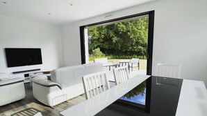 A modern living room with a flat-screen TV, white furniture, a dining table, and a large window with a view of greenery. - New house near Port-Manec'h beach (Nevez)