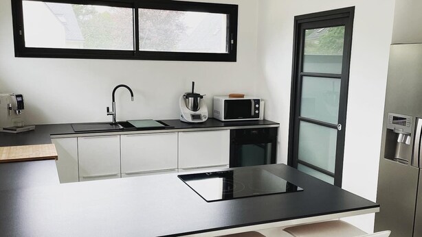 A modern kitchen with a black countertop, stainless steel appliances, a window above the sink, and a refrigerator on the right. - New house near Port-Manec'h beach (Nevez)