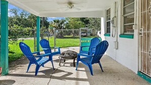 A covered patio with two blue chairs and a small table, a ceiling fan, and a view of a green lawn and house. - The Funky Cactus House - Team Joseph Ellen (Jacksonville)