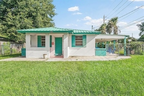 A small, single-story building with a green roof and white walls, a covered porch, and a fenced yard. - The Funky Cactus House | Team Joseph Ellen (Jacksonville)