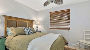 A bedroom with a bed, a nightstand, a lamp, a ceiling fan, a window with blinds, and a chair. - The Funky Cactus House - Team Joseph Ellen (Jacksonville)