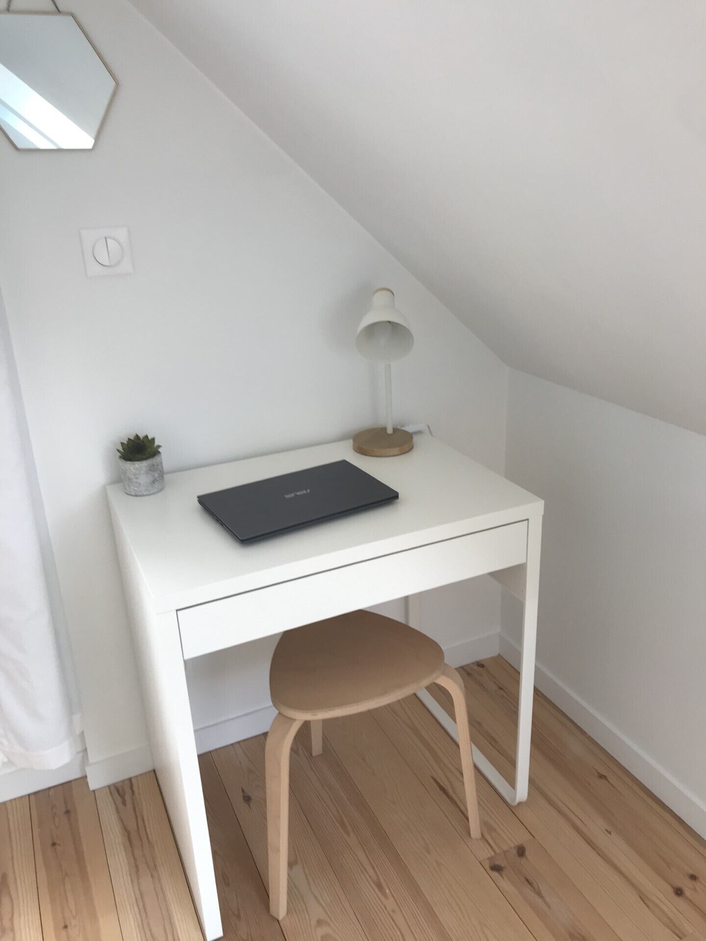 A white desk with a laptop, a small plant, and a desk lamp in a corner of a room with wooden flooring.