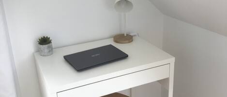 A white desk with a laptop, a small plant, and a desk lamp in a corner of a room with wooden flooring.