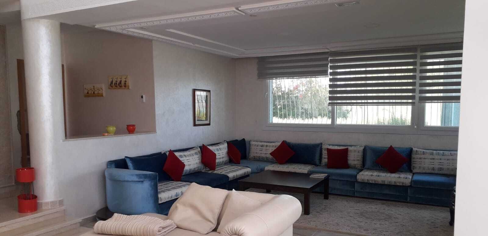 Una sala de estar con un sofá azul, una mesa de centro y un sillón color beige. Hay dos ventanas con persianas, un cuadro en la pared y un jarrón rojo y amarillo en una estantería.