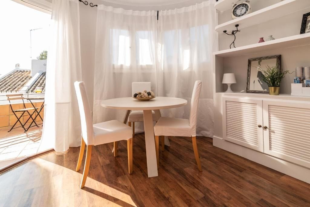 Un comedor con una mesa redonda y cuatro sillas, un armario blanco con un espejo y objetos decorativos, y vistas al exterior a través de cortinas traslúcidas.