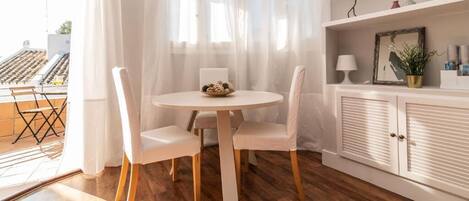 Un comedor con una mesa redonda y cuatro sillas, un armario blanco con un espejo y objetos decorativos, y vistas al exterior a través de cortinas traslúcidas.