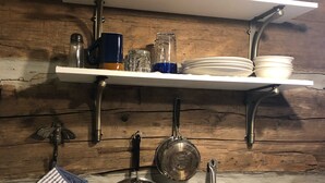 Una cocina con paredes de madera, una estufa blanca, ollas colgantes, un paño de cocina azul y blanco a rayas, y una repisa con platos y una vela.