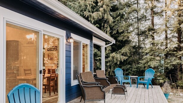 A wooden deck with outdoor seating, including chairs and a table, and a building with large windows and a porch. - 47MF - WiFi - Fireplace - Sleeps 6 3 Bedroom Home by RedAwning (Maple Falls)