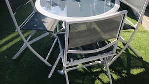 Une table ronde en verre, entourée de quatre chaises en métal, et un petit jardin avec du gazon artificiel et des plantes en pot.
