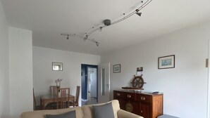 Un salon avec un canapé beige, une table basse en bois et une petite plante en pot. On y trouve également un meuble en bois orné d’un motif de roue de navire et un tableau encadré accroché au mur.