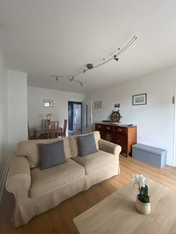 A living room with a beige sofa, a wooden coffee table, and a small potted plant. There is a wooden cabinet with a ship's wheel decoration and a framed picture on the wall.