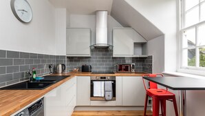A modern kitchen with a built-in dishwasher, a microwave, and a kettle on the counter. There is a window providing natural light and a red chair at the dining table. - 14 Feversham House · Stylish 1-Bed Apartment with York Minster Views - Pass The Keys (York)