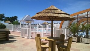 An outdoor patio with wooden tables and chairs, a pool area with a white fence, and thatched umbrellas. - Lovely mobile home in a 4-star residential estate in Les Sables D'Olonne (Les Sables-d'Olonne)