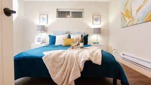 A bedroom with a large bed, two bedside tables, a window with blinds, and a wall-mounted heater. - Burnaby Cozy suite Near Skytrain (Burnaby)