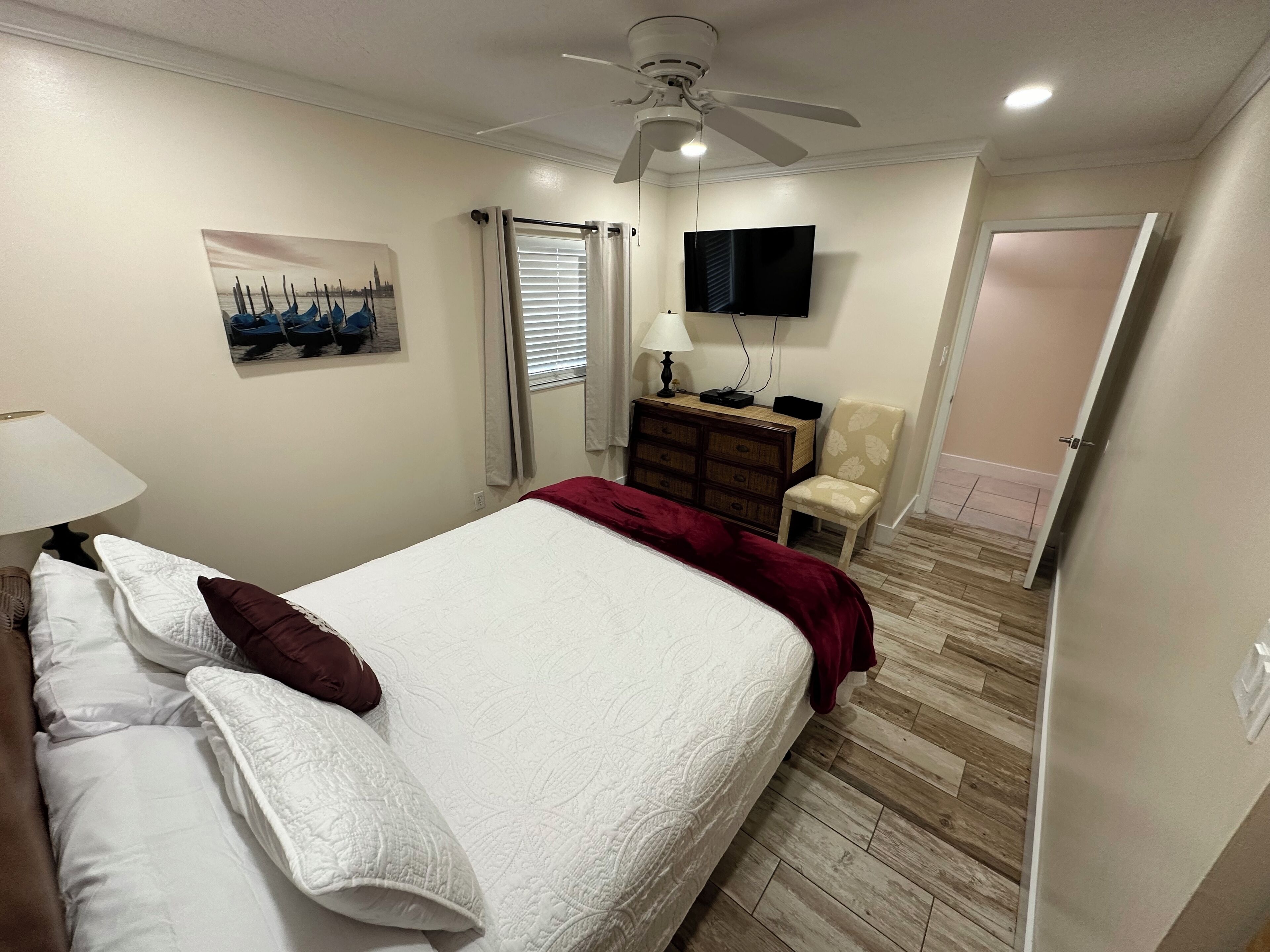 Un lit bien fait, avec une literie blanche et bordeaux, un ventilateur de plafond, un téléviseur fixé au mur, une commode en bois et une chaise.