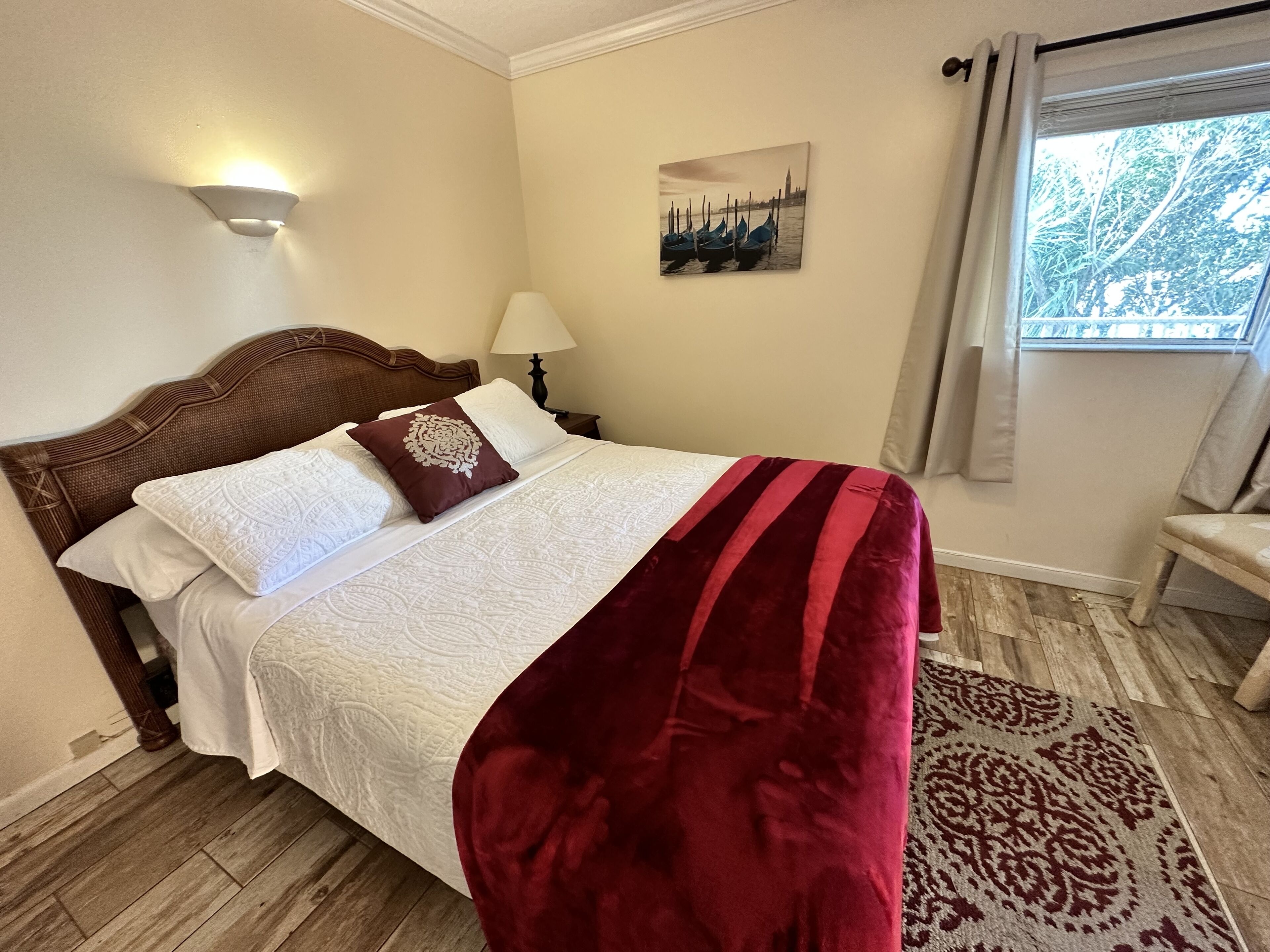 A neatly made bed with a white duvet, a brown decorative pillow, and a red blanket. There is a lamp on a bedside table, a painting of a cityscape on the wall, and a window with curtains.