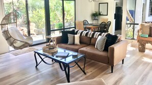 A spacious living room with a large window, a hanging chair, a brown sofa with striped cushions, and a glass coffee table. - Casa del Sol - Peaceful home away from home. (Santa Barbara)
