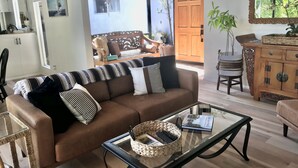 A living room with a brown sofa, a glass coffee table, and a wicker basket. There is a wooden door and a window with a view of greenery outside. - Casa del Sol - Peaceful home away from home. (Santa Barbara)