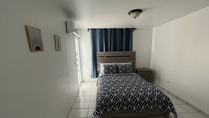 Un dormitorio con cama, mesita de noche, aire acondicionado instalado en la pared y ventana con cortinas.