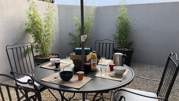 Une table ronde d’extérieur, entourée de chaises en métal et de plantes en pot, placée sous un grand parasol.