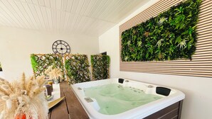 A hot tub with a wooden deck, surrounded by lush green plants and a decorative clock on the wall. - Villa with spa pool petanque court (Narbonne)