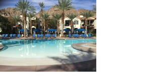 A pool area with lounge chairs and umbrellas, surrounded by palm trees and buildings with Mediterranean architecture. - New Listing - Lovely Mountain View Studio for two (La Quinta)