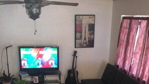 Ett vardagsrum med en tv, en takfläkt, en gitarr, en soffa och två fåtöljer.
