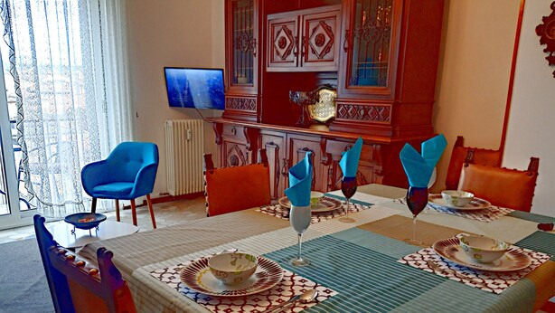 Zona pranzo con tavolo apparecchiato per quattro, sedie, televisore e mobiletto decorativo.