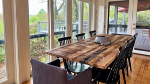 Een houten eettafel met stoelen, grote ramen en uitzicht op groen buiten.