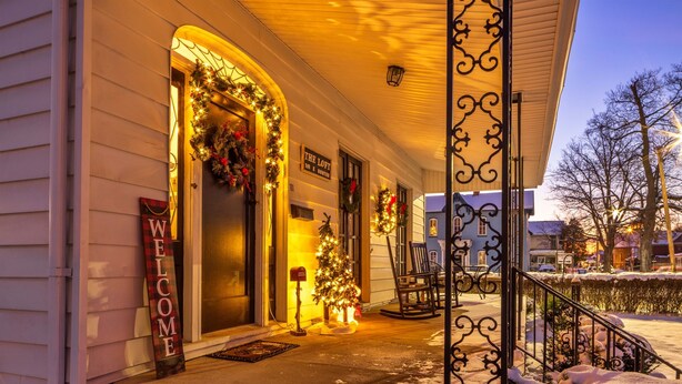 Una casa con portico decorato con luci natalizie e ghirlande, un cartello di benvenuto e un piccolo abete decorato.