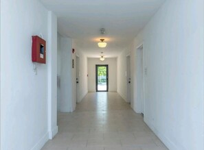 A long, narrow hallway with white walls and a tiled floor, leading to a door at the end.