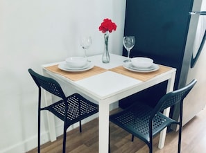 A white dining table with two black chairs, a vase with red flowers, and two wine glasses on the table.