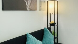 En sort sofa med to turkise puder, et indrammet portræt af en tiger, en vægmonteret lampe og en lille plante på et sort metalstativ.