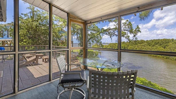 A covered outdoor area with a glass table and chairs, overlooking a body of water and greenery.