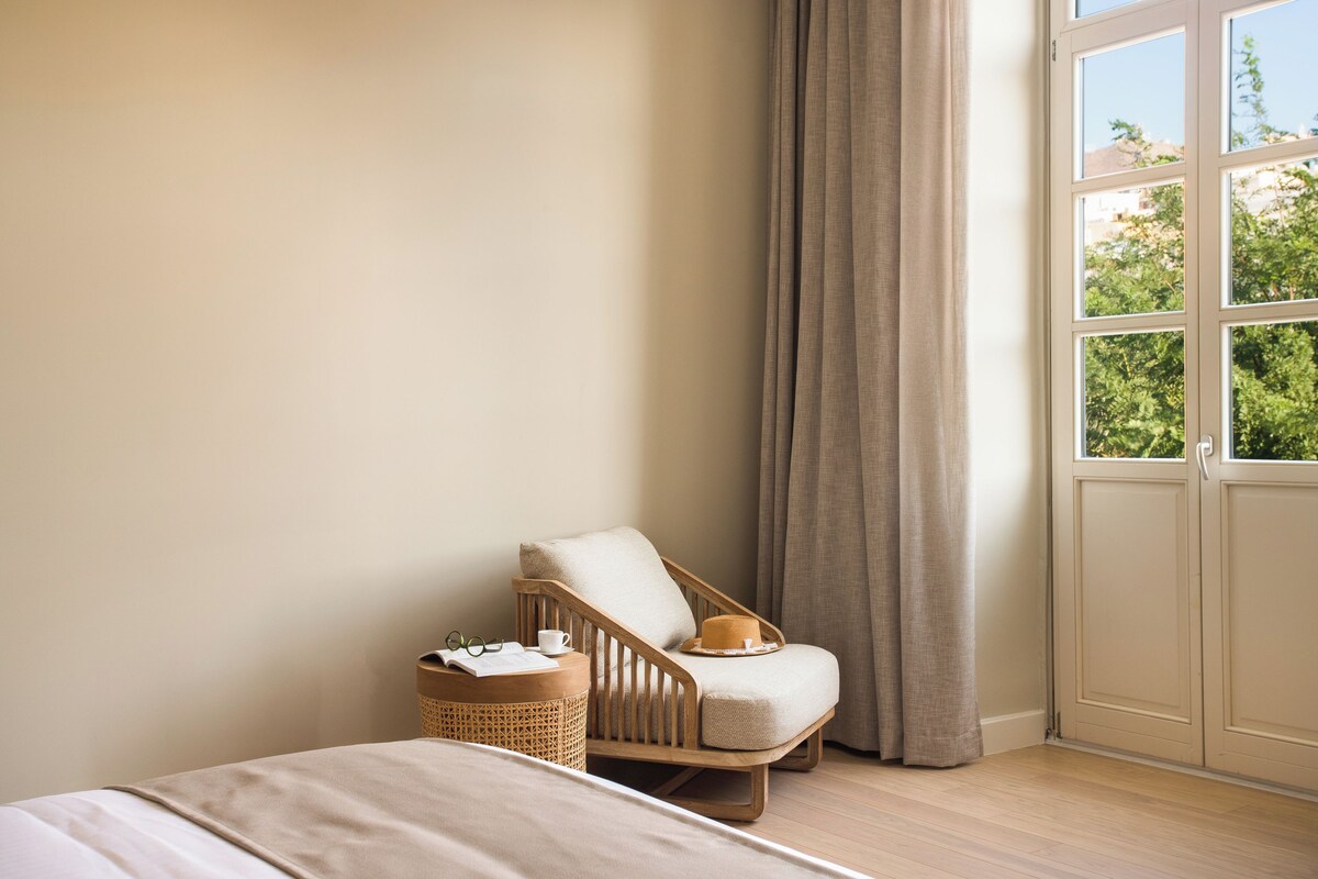 A bedroom with a bed, a wooden chair, a wicker basket, and a window with curtains.
