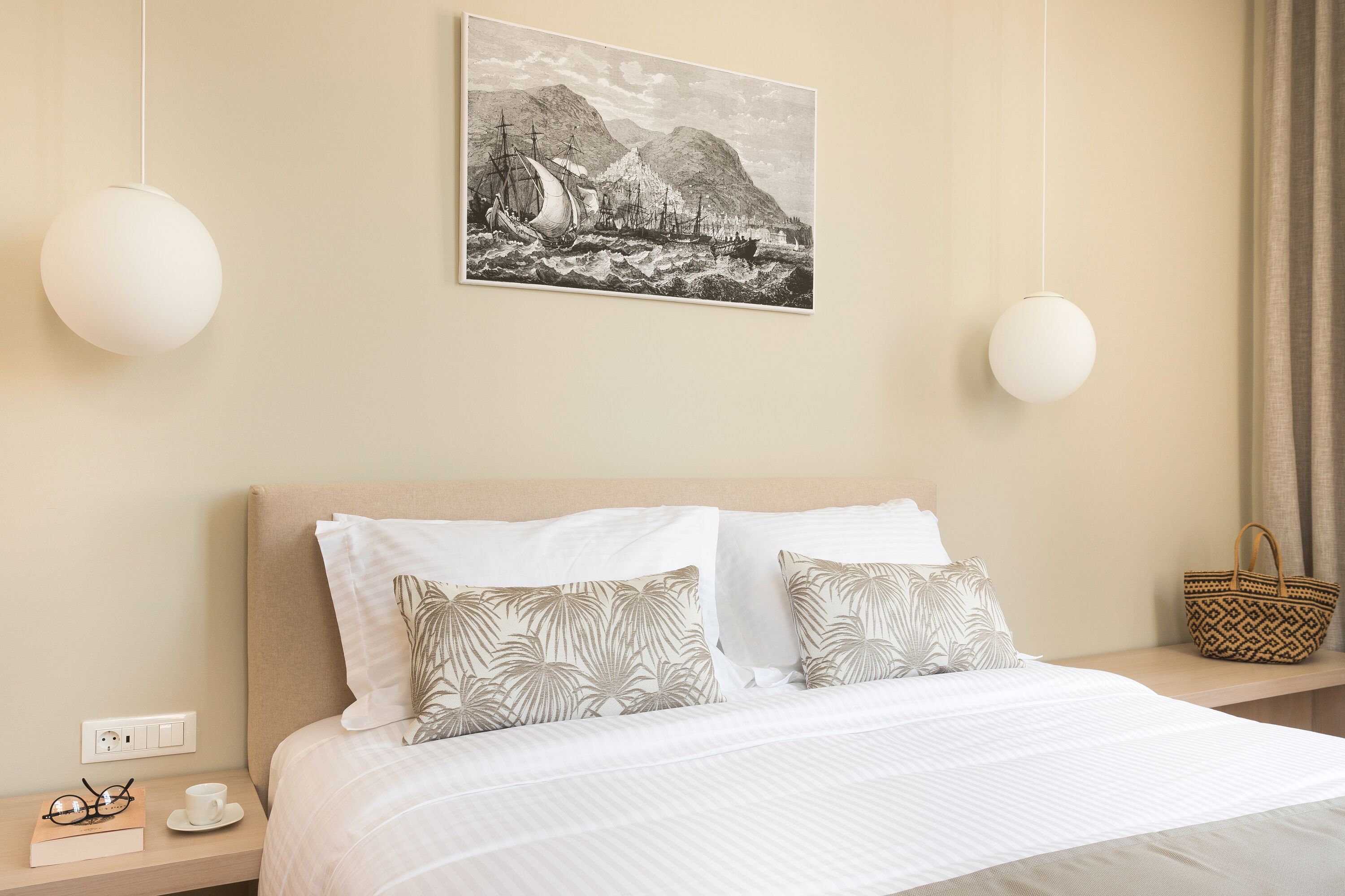 A neatly arranged hotel room with a bed, pillows, a bedside table with a book and glasses, a wall-mounted painting, and a hanging light fixture.