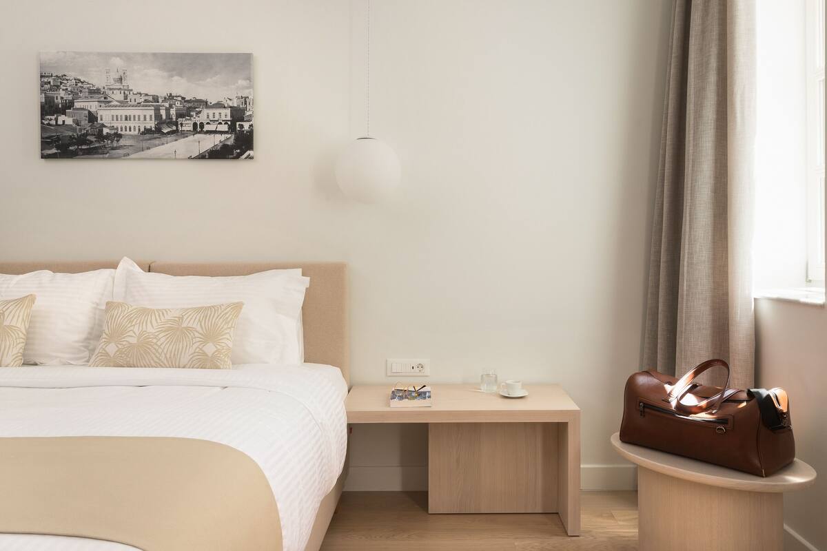 A hotel room with a bed, bedside table, lamp, and a handbag on a side table.