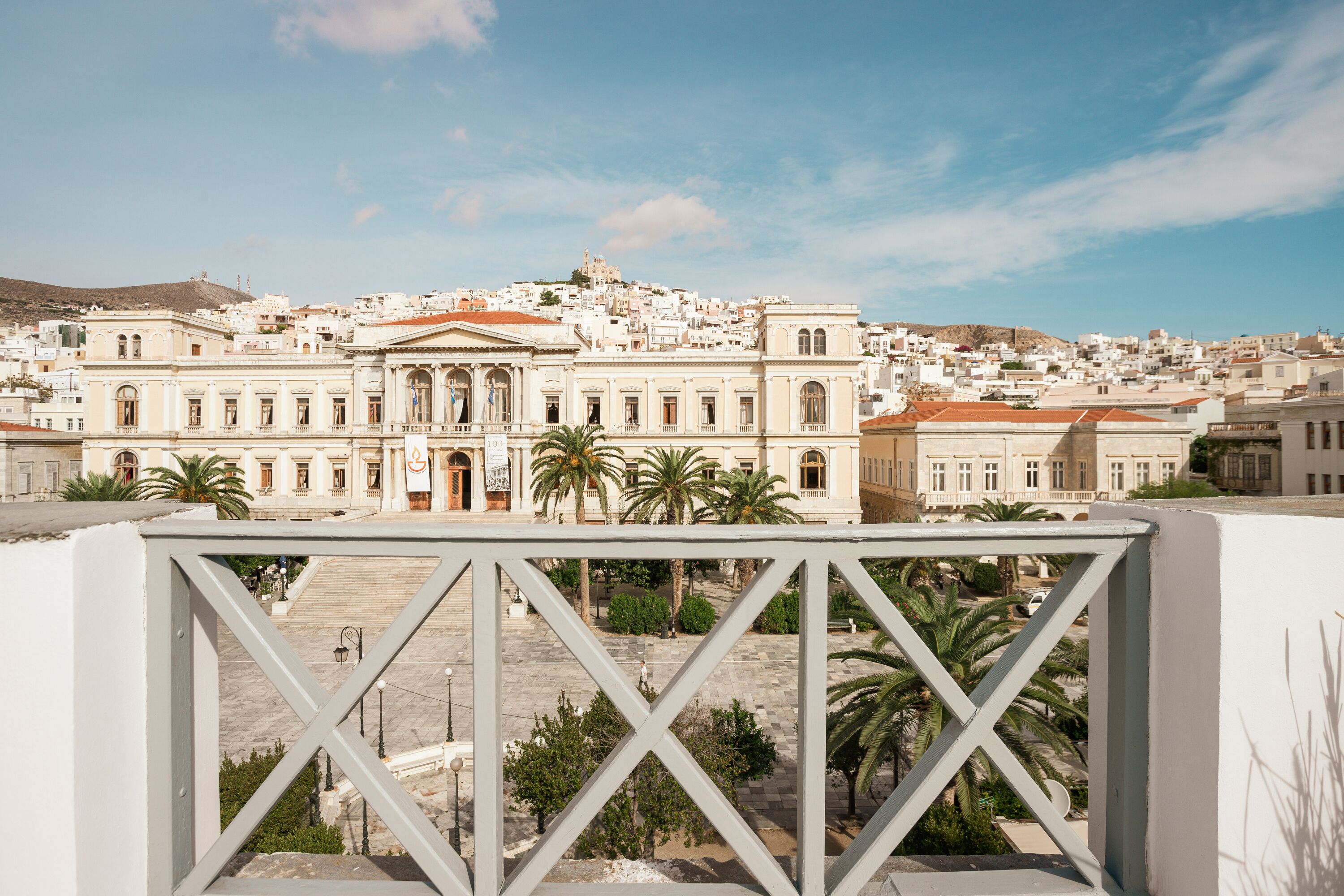 A view of a historic building with classical architecture, a courtyard with palm trees, and a clear sky.