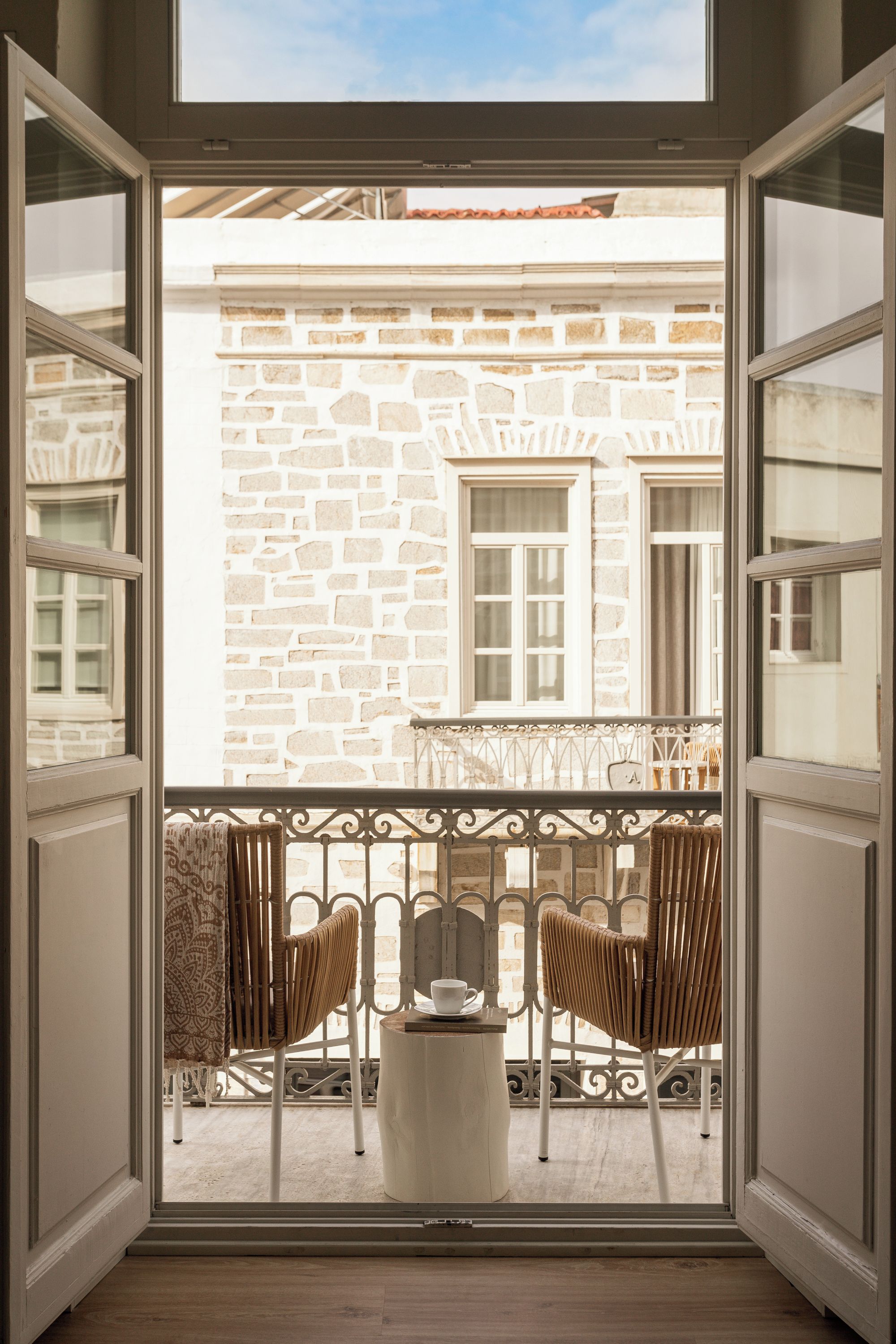 Un balcon ouvert avec un mur en pierre, deux fauteuils en osier, une petite table et une fenêtre donnant sur un immeuble.