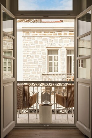 An open balcony with a stone wall, two wicker chairs, a small table, and a window with a view of a building.