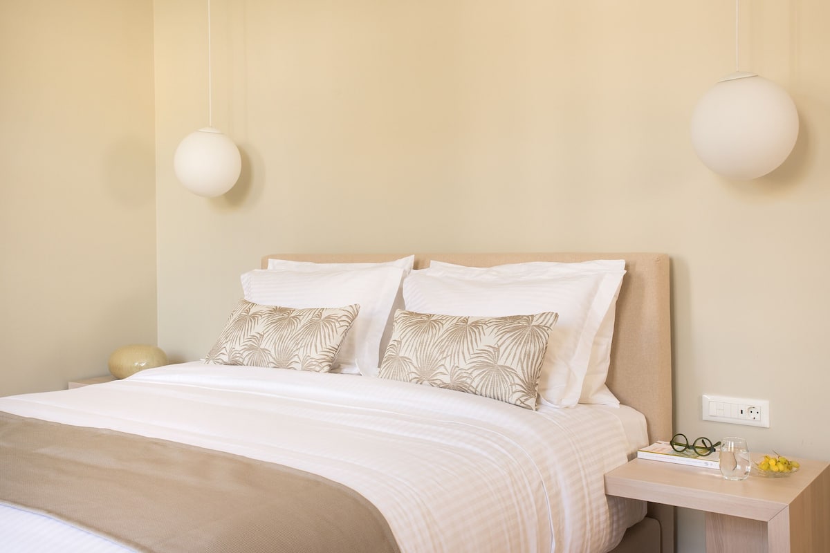 A neatly made bed with a beige headboard, two pillows, and a bedside table with a lamp and a book.