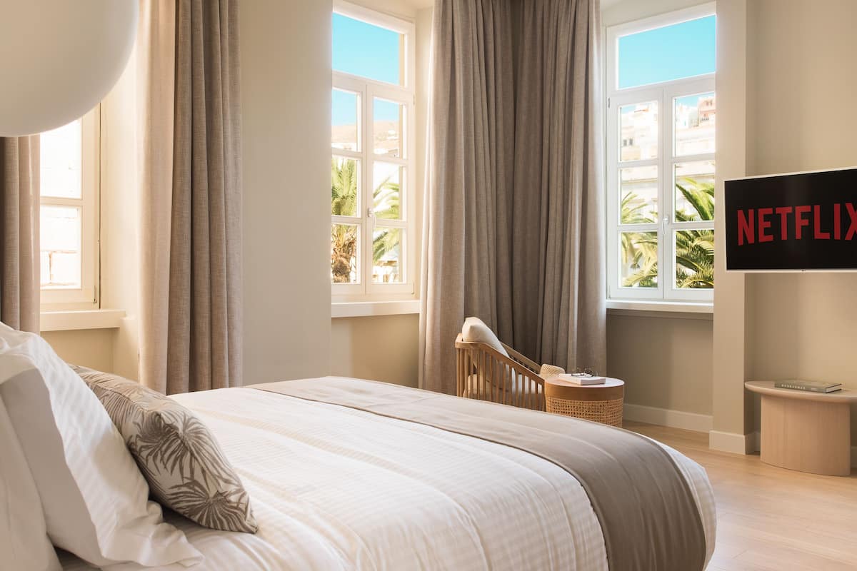 A bedroom with a bed, curtains, a TV, and a view of palm trees.