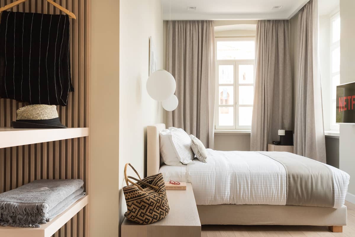 A neatly arranged bedroom with a bed, a bedside table with a lamp, a hanging coat rack, and a window with curtains.