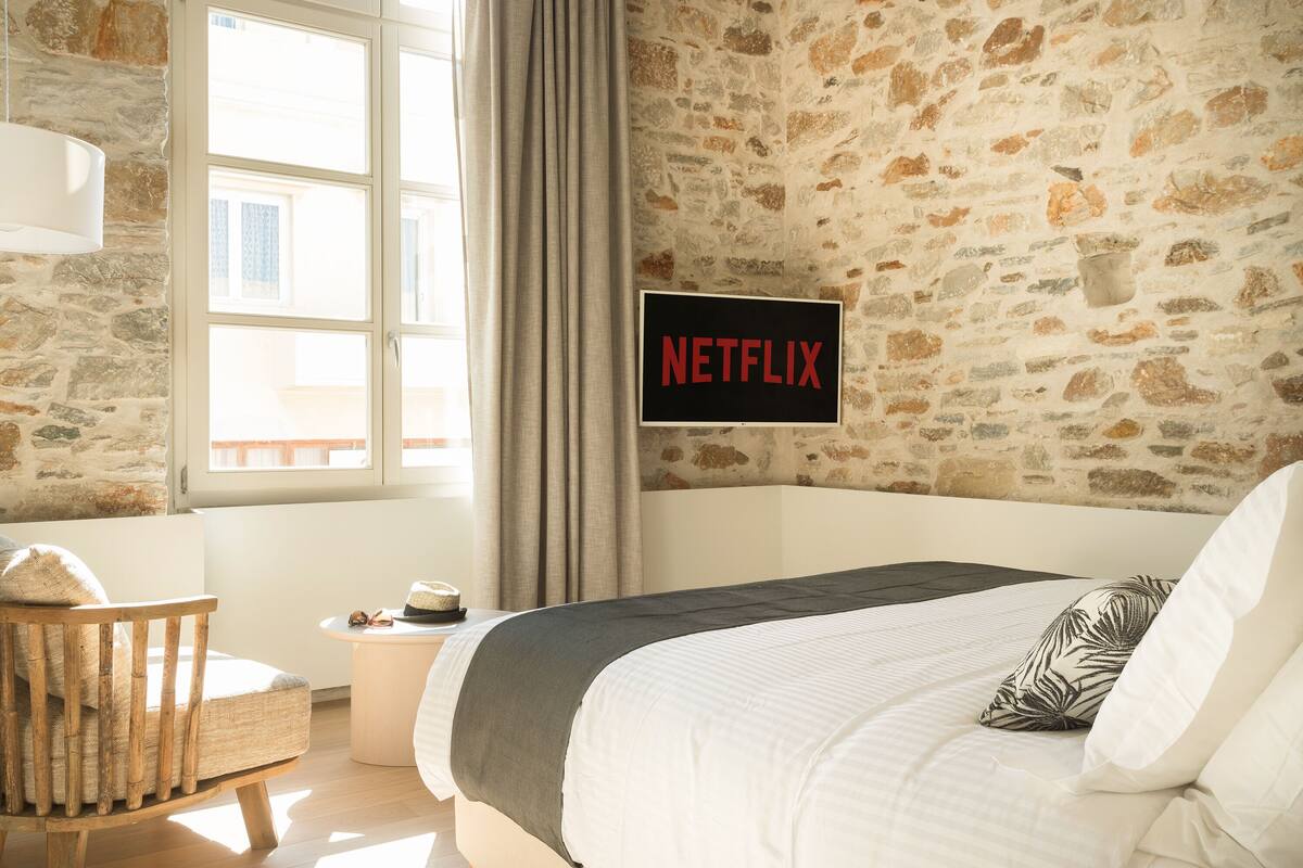 A bedroom with a stone wall, a bed with a white comforter, a wooden chair, a small table with a hat and sunglasses, and a sign that says "NETFLIX".