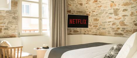 Un dormitorio con pared de piedra, una cama con cubrecama blanco, una silla de madera, una mesita con un sombrero y gafas de sol, y un cartel que dice "NETFLIX".