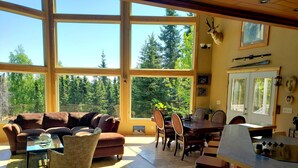 A cozy living room with a sofa, chairs, a dining table, and a kitchen area with a bar. Large windows offer a view of the forest. - Windows With a View (Soldotna)