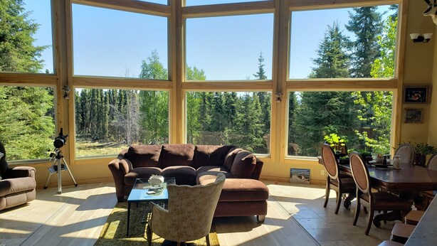 A spacious living room with large windows, a brown sofa, a coffee table, and a dining area with chairs and a table. - Windows With a View (Soldotna)