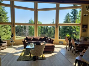 A spacious living room with large windows, a brown sofa, a coffee table, and a dining area with chairs and a table. - Windows With a View (Soldotna)