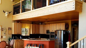 A spacious living area with a high ceiling, a kitchen with modern appliances, and a visible staircase leading to an upper level. - Windows With a View (Soldotna)